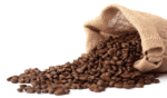 A burlap sack labeled "Café Rico Cafe Mundo Coffee Beans, 100% Arabica" is tipped over, spilling a pile of dark brown and glossy roasted coffee beans onto a white surface. These beans embody the aroma cherished by every café's barista. The texture of the burlap bag is coarse and rustic, adding to its charm.