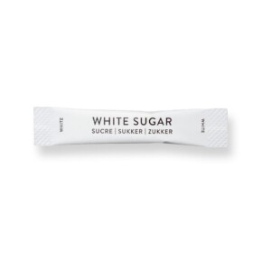 A small white packet labeled "WHITE SUGAR" in bold black letters sits ready for a café moment, with the words "SUCRE," "SUKKER," and "ZUKKER" beneath. The ends of the packet also display the word "WHITE." A barista's touch completes the plain white backdrop.
