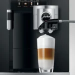 The Jura GIGA X8 Pro Gen II Black Tanked Bean to Cup Coffee Machine, with its advanced technology, creates a layered cappuccino in a glass, reminiscent of a skilled barista's touch. Its café-style digital interface showcases various drink options, and the coffee features a distinct foam layer on top.