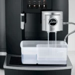 The Jura GIGA X8 Pro Gen II Black Tanked Bean to Cup Coffee Machine stands like a skilled barista, expertly dispensing liquids into a transparent container. This café-worthy machine features a digital display showcasing various drink options.