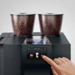 Channeling their inner barista, a person selects a coffee option on the touchscreen of the sleek Jura GIGA X8 Pro Gen II Black Tanked Bean to Cup Coffee Machine, adorned with two transparent containers filled with aromatic beans, bringing the essence of a chic café into their own space.