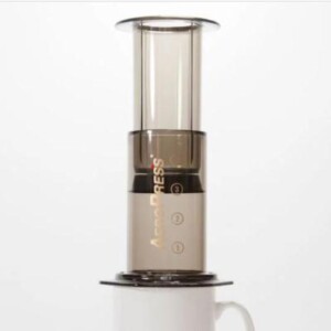 AeroPress coffee maker which combines affordability and simplicity with the ability to produce top quality coffee, the aeropress sits on a white porcelain cup and coffee is slowly dripping through the filter.